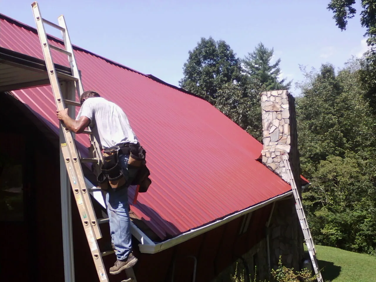 Roofer on a ladder beside a red metal roof and stone chimney — Soffit & Fascia Repair by Roofing Brothers in Clarissa